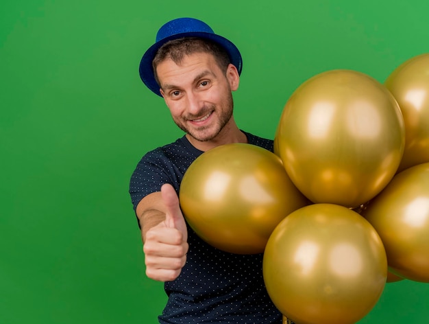 Smiling handsome man wearing blue party hat holds helium balloons and thumbs up isolated on green wall with copy space