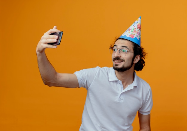 眼鏡と誕生日の帽子を身に着けているハンサムな男の笑顔は、オレンジ色の背景で隔離の自撮り