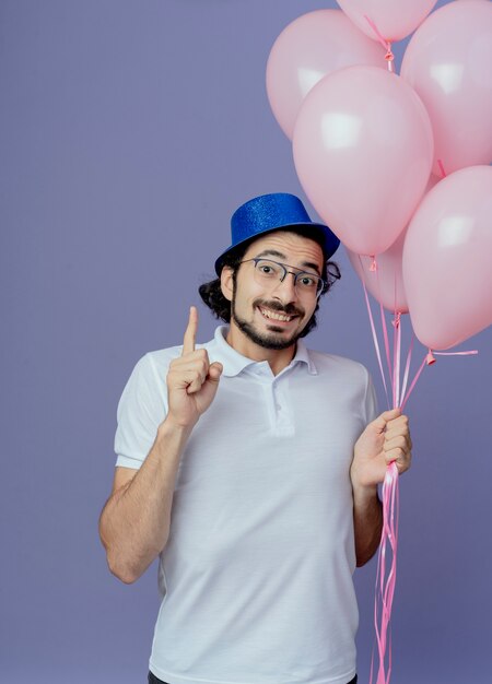 Smiling handsome man wearing glasses and blue hat holding balloons and points at up isolated on purple