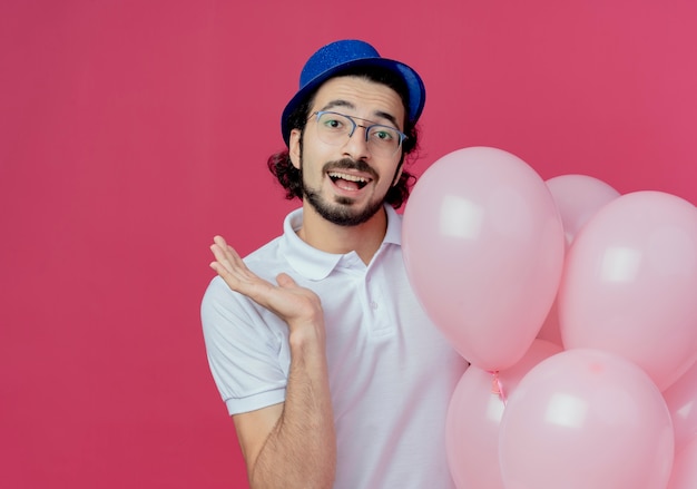 Smiling handsome man wearing glasses and blue hat holding balloons and points with hand to side isolated on pink background with copy space