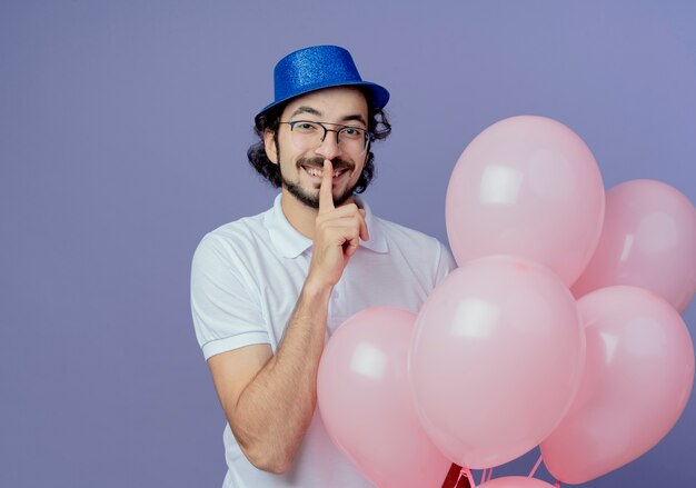 Smiling handsome man wearing glasses and blue hat holding balloons and showing silence gesture isolated on purple