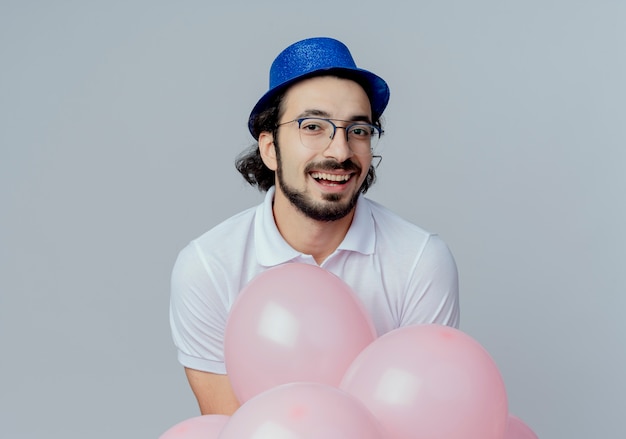 Uomo bello sorridente con gli occhiali e cappello blu in piedi dietro palloncini isolati su bianco