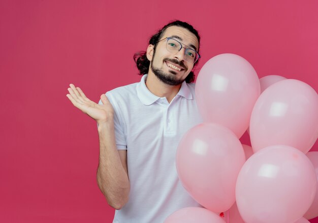 Smiling handsome man wearing glasses holding balloons and points with hand to side isolated on pink background with copy space