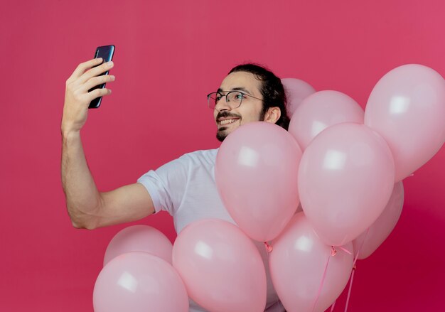 Улыбающийся красавец в очках держит воздушные шары и делает селфи на розовом
