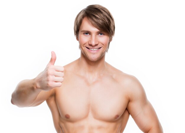 Smiling handsome man with muscular torso shows thumbs up sign - isolated on white wall.