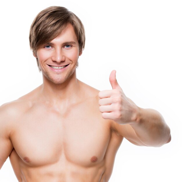 Smiling handsome man with muscular torso shows thumbs up sign - isolated on white wall.