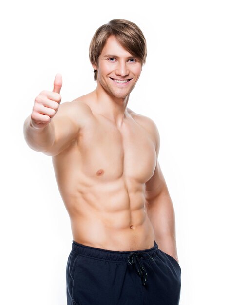 Smiling handsome man with muscular torso shows thumbs up sign - isolated on white wall.