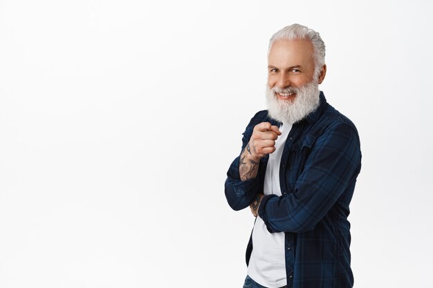 Smiling handsome senior man with long beard, pointing finger at front and looking happy, choosing, inviting or recruiting people, praising person, standing over white wall