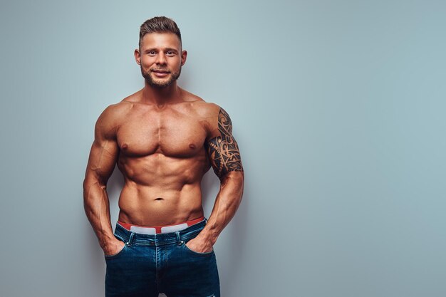 Smiling handsome shirtless bodybuilder with stylish haircut and beard, with tattoo on his arm, posing in a studio. Isolated on a gray background.