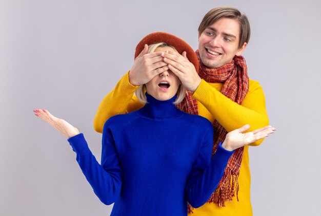 Smiling handsome slavic man with scarf around his neck closing eyes of surprised pretty blonde woman with beret on isolated on white wall with copy space