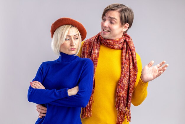 Smiling handsome slavic man with scarf around his neck looking at offended pretty blonde woman with beret standing with crossed arms isolated on white wall with copy space