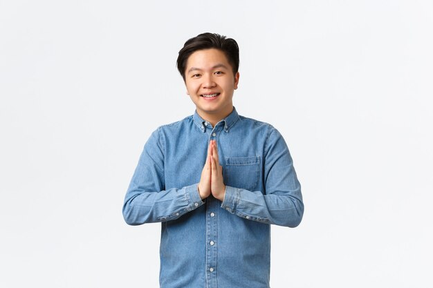 Smiling handsome young asian man looking thankful and pleased, holding hands together to bow politely, greeting someone, say namaste, standing white background delighted