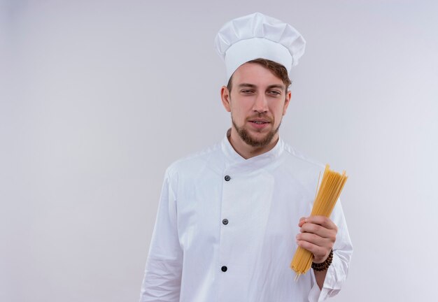 Un sorridente bello giovane barbuto chef uomo che indossa l'uniforme bianca del fornello e cappello che tiene gli spaghetti mentre guarda un muro bianco