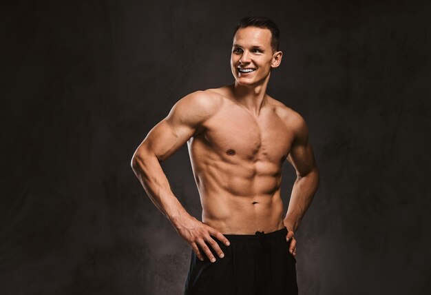 Smiling handsome young fitness model with muscular body posing in studio on a dark background.