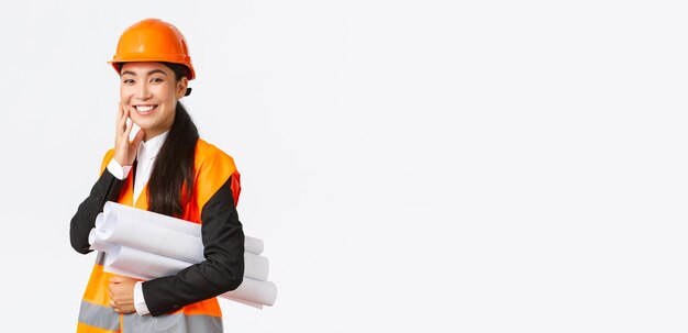 Smiling happy asian female architect construction manager in safety helmet and jacket carry blueprints of building project and looking delighted finish plan in time standing white background