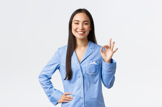 Smiling happy asian girl in blue jammies showing okay gesture in like or support, say OK as recommend great quality product, guarantee all under control, saying everything good, white background