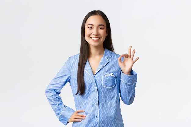 Улыбающаяся счастливая азиатская девушка в синих джемперах показывает нормальный жест в знак лайка или поддержки, говорит ОК, рекомендует продукт отличного качества, гарантирует, что все под контролем, говорит все хорошо, белый фон