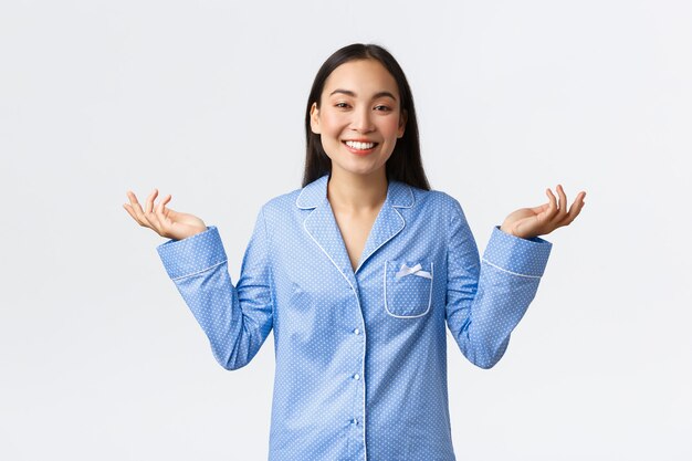 Smiling happy asian girl in jammies raising hands up shrugging, dont know anything, have no clue. uncertain or clueless woman in pajamas looking unbothered over white background