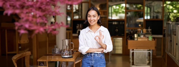 笑顔の幸せなアジアの女性起業家が、OK OKサインインの承認を示すカフェを勧める