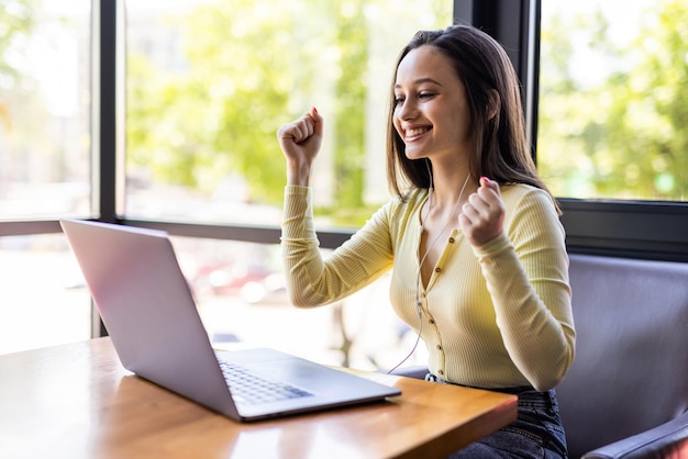 Smiling happy businesswoman in headphones sits at desk, looks at laptop screen, making notes, participating in self-improvement webinar