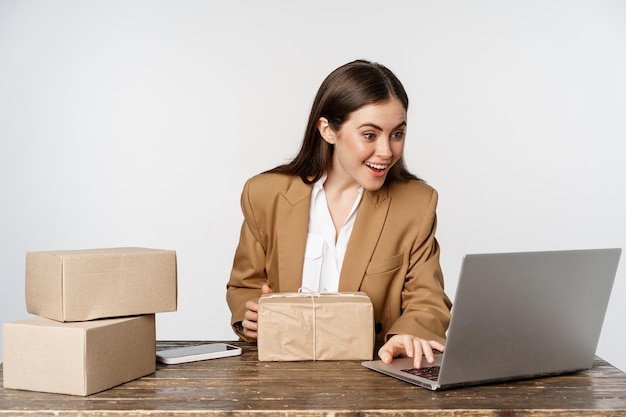 Donna d'affari felice sorridente che lavora nel suo ufficio con ordini di processo di consegna pacchi sul suo laptop...