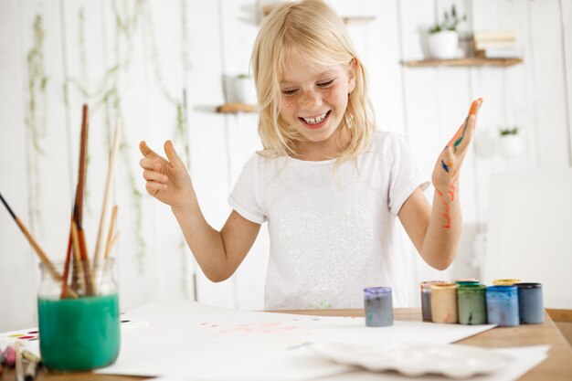 Smiling, happy and cheerful blonde girl showing her teeth, having fun while painting. Female freckled child messed up her hand with paint of different colors.