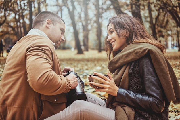 Smiling happy couple is drinking hot drink from thermos while sitting at autumn park.