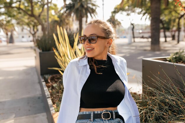 Smiling happy europnean woman in sunglasses is resting on sunny street with palms and looking aside Portrait of model woman with smile on face holds hat with hands and wears sunglasses