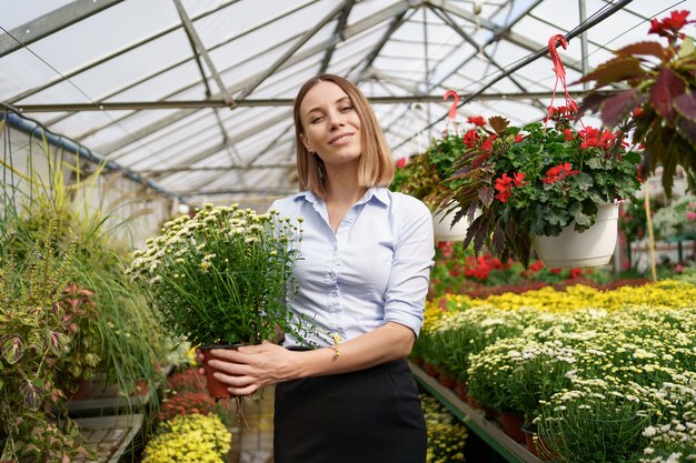 彼女は温室の園芸植物に傾向があるように彼女の手で鉢植えの菊を持って立っている彼女の保育園で幸せな花屋を笑顔