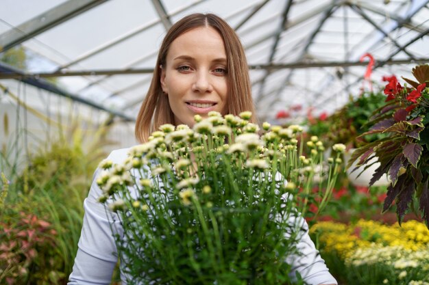 그녀는 온실의 정원 식물에 경향이 그녀의 손에 화분에 심은 국화를 들고 그녀의 보육 서에서 웃는 행복 꽃집