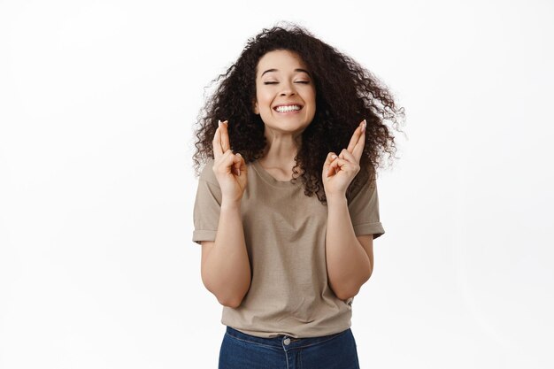 Smiling happy girl makes wish, hopeful cross fingers, wishing for good luck, close eyes with excited face expression, standing over white background