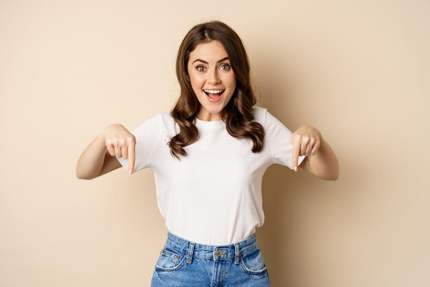 Smiling happy girl pointing fingers down and showing banner, company logo below, click link, standing over beige background.