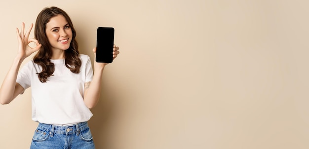 Улыбающаяся счастливая девушка показывает приложение для экрана мобильного телефона на смартфоне и хорошо рекомендует приложение для покупок в интернет-магазине на бежевом фоне
