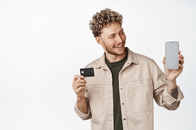 Smiling happy guy shows credit card and mobile phone screen looks at application with satisfied face expression white background