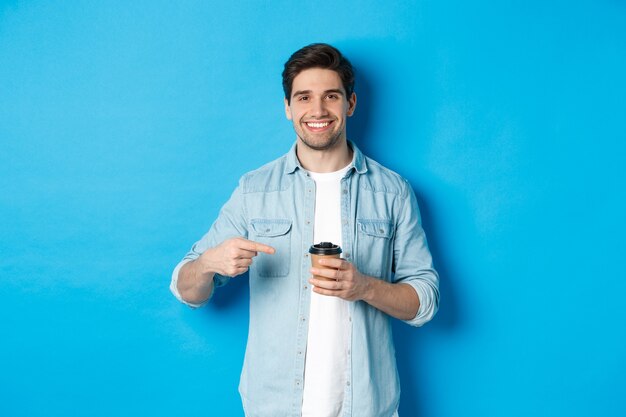 Smiling happy man pointing at paper cup with coffee, recommending cafe, standing over blue background