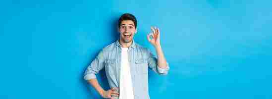 Free photo smiling happy man showing ok sign and looking pleased approving something good standing against blue