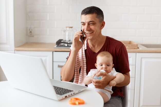 Улыбающийся счастливый человек в бордовой повседневной футболке с полотенцем на плече, присматривает за ребенком и работает в сети из дома, имеет приятный разговор с клиентом или партнером.