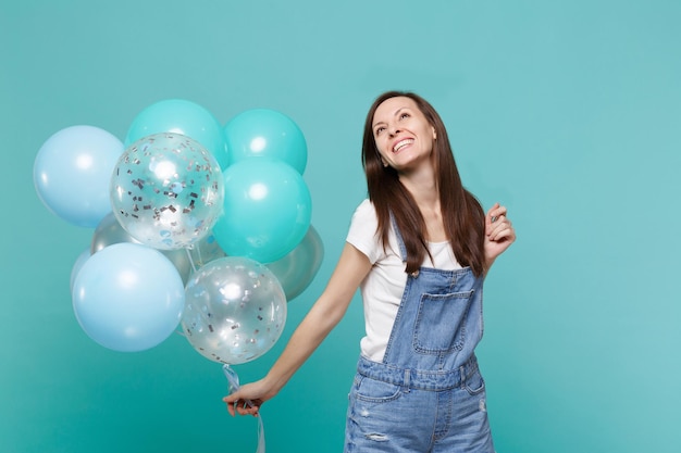 Smiling happy pensive young woman in denim clothes looking up celebrating and holding colorful air balloons isolated on blue turquoise wall background. birthday holiday party, people emotions concept.