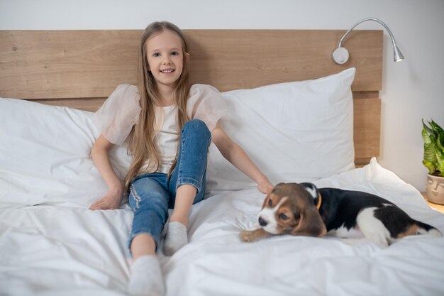 웃고 있는 행복한 예쁜 소녀가 침대에 앉아 차분한 귀여운 강아지를 쓰다듬어 줍니다