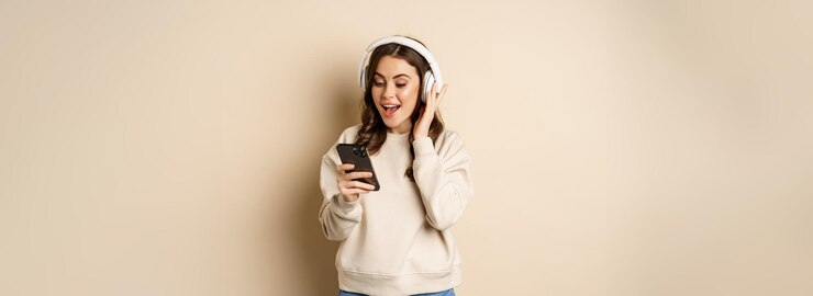 無料Photo ベージュ色の背景の上に立って興奮して笑っているスマートフォンでヘッドフォンで音楽を聴いて笑顔の幸せな女性