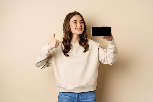 Smiling happy woman showing smartphone horizontal screen, thumbs up, recommending website, online store or app, standing over beige background