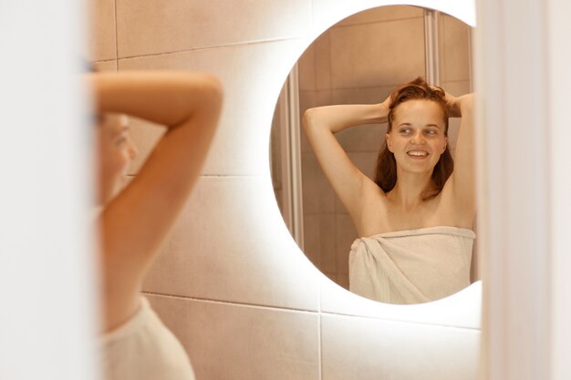 腕を上げたバスルーム、朝のルーチン、魅力的な女性の包まれた白いタオルの鏡の前に立っている完璧な肌を持つ笑顔の幸せな女性。