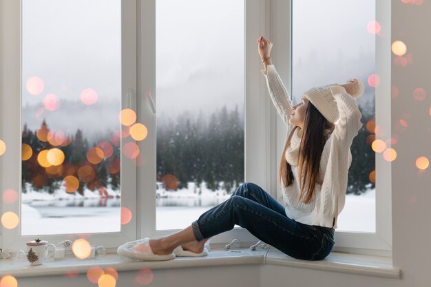 Smiling happy young attractive woman in stylish white knitted sweater, scarf and hat sitting at home on windowsill at Christmas having fun holding hands up, winter forest background view, lights bokeh