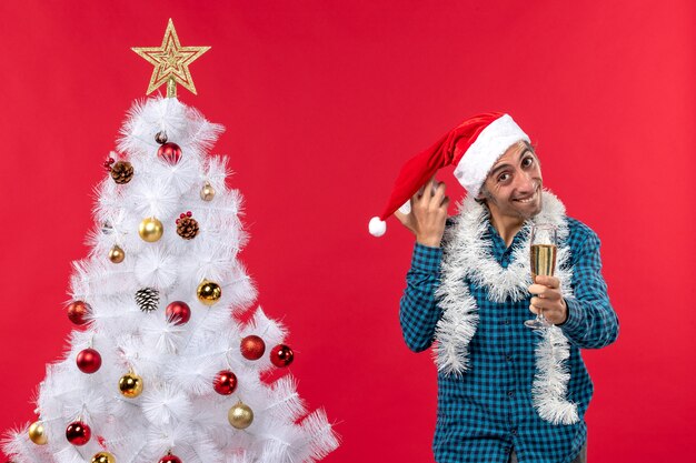 행복 한 젊은 남자 산타 클로스 모자와 함께 연주 하 고 와인 한 잔을 제기 하 고 빨간색에 크리스마스 트리 근처에 서 시계를 들고 웃 고
