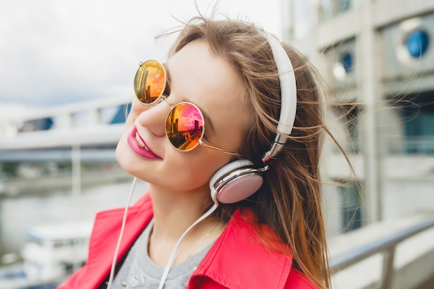 Smiling happy young hipster woman in pink coat listening to music on headphones, wearing sunglasses