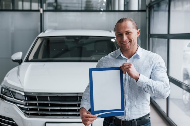 Smiling and having good mood. Manager stands in front of modern white car with paper and documents in hands