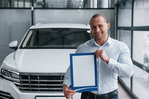 Улыбка и хорошее настроение. Менеджер стоит перед современной белой машиной с бумагами и документами в руках