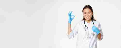 무료 사진 고무 장갑과 의료 유니폼에 웃는 의료 노동자 아시아 여자 의사는 승인 오카를 보여줍니다