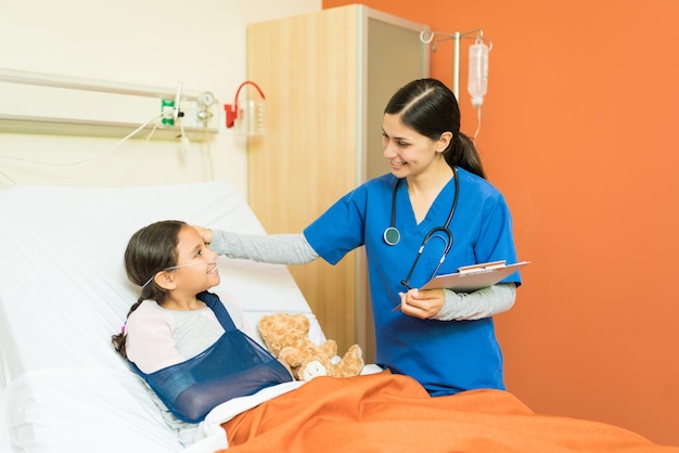 Улыбающийся медицинский работник держит отчеты, глядя на маленького пациента с переломом руки, лежащего в больнице