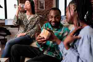 Free photo smiling heartily man offering gift to woman friend while enjoying wine party together at home. group of happy friends in living room celebrating birthday while exchanging presents and drinking wine.